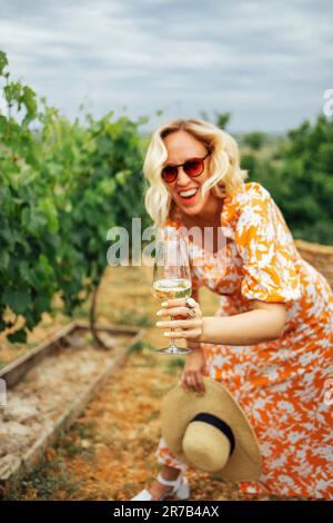 Allegra donna bionda in abito arancione brillante con stampa floreale tiene un bicchiere di champagne sopra la testa in vigna. Bella ragazza in occhiali da sole lau Foto Stock