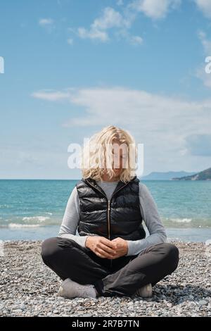 Adulto turista europeo donna bionda seduta da sola su una spiaggia di ciottoli con oceano blu e cielo sullo sfondo, ripresa verticale. Persone e vacanze estive Foto Stock