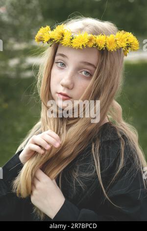 Una ragazza seria e misteriosa in una corona di dente di leone. Una ragazza con lunghi capelli spessi è triste in primavera per strada. Foto Stock