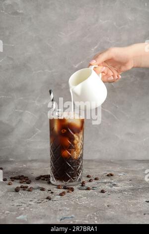 Donna che versa il latte dalla brocca in un bicchiere di caffè ghiacciato con paglia e fagioli sul tavolo grunge grigio vicino alla parete Foto Stock