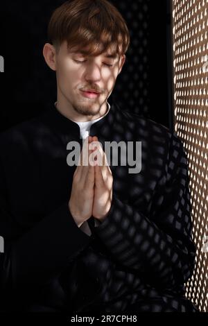 Sacerdote cattolico che prega vicino alla finestra di legno in cabina confessionale Foto Stock