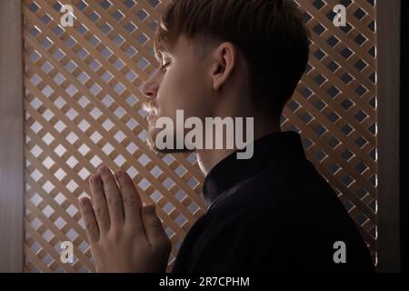 Sacerdote cattolico che prega vicino alla finestra di legno in cabina confessionale Foto Stock