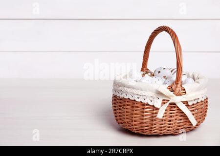 Cestino di vimini con uova di Pasqua decorate in modo festivo su un tavolo di legno bianco. Spazio per il testo Foto Stock