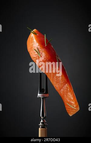 Salmone affumicato su una forchetta. Pezzo di pesce con rosmarino su sfondo nero. Foto Stock