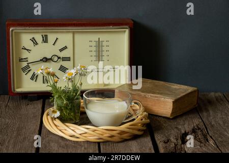 una tazza di latte e un vaso con margherite vicino all'orologio con un libro su un tavolo di legno Foto Stock