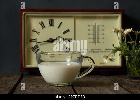 una tazza di latte e un vaso con margherite vicino all'orologio su un tavolo di legno Foto Stock