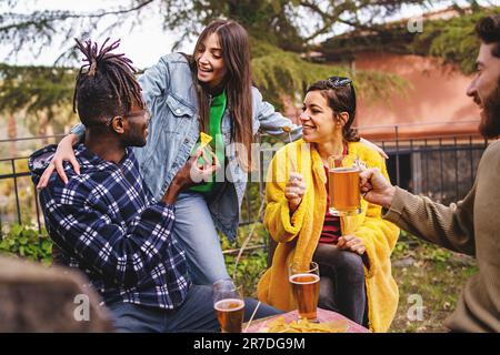 Un gruppo diversificato di amici abbraccia in una birreria all'aperto, gustando birra, nachos e antipasti di olive. Foto Stock