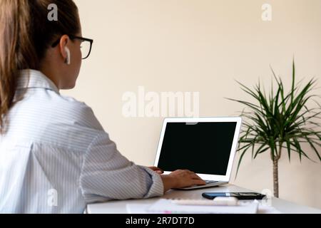 La donna d'affari con vista posteriore indossa occhiali e auricolari wireless che lavorano sul suo computer portatile in ufficio, schermo vuoto del computer portatile. Foto Stock