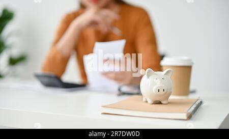 Immagine ravvicinata di una banca di piggy su un tavolo con una donna che pianifica le sue spese mensili dietro. spese, risparmio, bilancio, reddito, prestito della casa. i sfocato Foto Stock