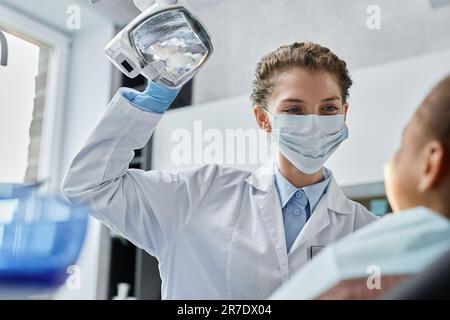 Ritratto di amichevole dentista donna indossare maschera e regolare le luci mentre si lavora con il bambino in clinica dentale, spazio copia Foto Stock