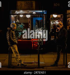 Due maschi adulti che si godono una pausa presso una fermata dell'autobus pubblico mentre utilizzano i loro dispositivi elettronici mobili Foto Stock