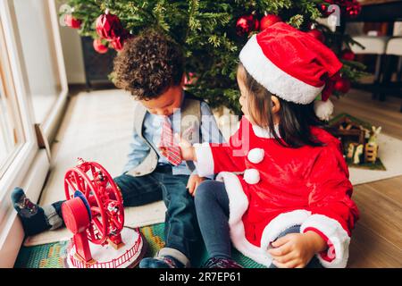 Gruppo di due adorabili bambini di 3 anni che giocano accanto all'albero di Natale. Bambina che ammira la cravatta del ragazzo Foto Stock