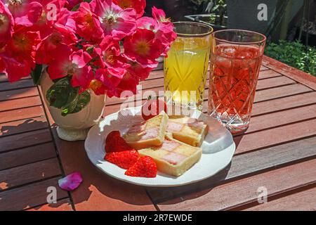 Un piatto di fettine di battenberg su un tavolo da giardino ardesiato con corone morbide e aposy di rose rampicanti; Foto Stock