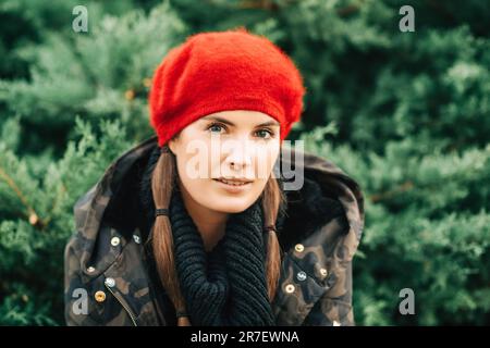 Ritratto in primo piano all'aperto di una bella giovane donna che posa su sfondo verde indossando giacca militare e berretto rosso Foto Stock