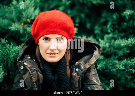 Ritratto in primo piano all'aperto di una bella giovane donna che posa su sfondo verde indossando giacca militare e berretto rosso Foto Stock