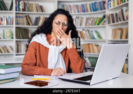 Giovane studente assonnato e stanco che studia nella biblioteca accademica del campus fino a tardi, donna ispanica che si prepara per gli esami di ingresso tra i libri, utilizzando il portatile per visualizzare i corsi online. Foto Stock