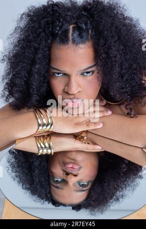 Una bella donna afroamericana che guarda con attenzione in uno specchio rotondo con le mani sul viso, seduta in una stanza elegantemente decorata Foto Stock