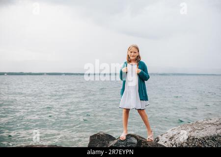 Ritratto all'aperto di adorabile bambina che gioca accanto al lago, indossando un abito bianco e una giacca verde cardigan Foto Stock