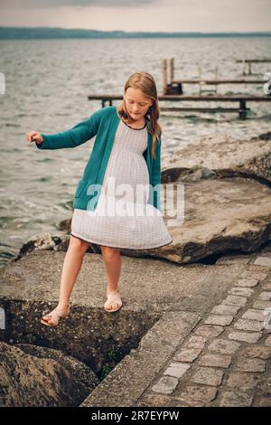 Ritratto all'aperto di adorabile bambina che gioca accanto al lago, indossando un abito bianco e una giacca verde cardigan Foto Stock