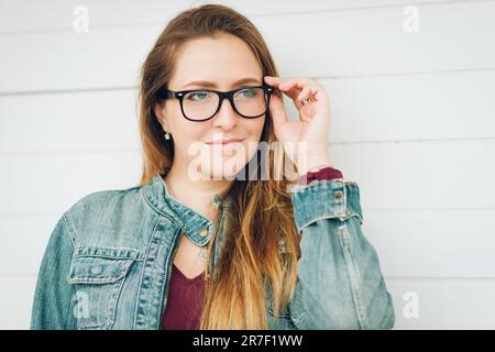 Ritratto all'aperto di giovane bella donna che indossa occhiali e giacca in denim, in posa su sfondo bianco Foto Stock