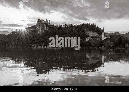 immagine in bianco e nero di un effetto solare mentre il sole tramonta per creare una splendida scena di castello di bled e chiesa di san martino riflessa nel lago di bled Foto Stock