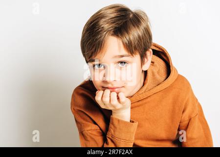 Studio di un bel ragazzo di 10 anni con capelli biondi, con cappuccio marrone, posato su sfondo bianco Foto Stock