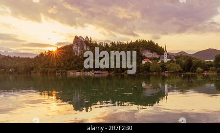 l'effetto sunburst mentre il sole tramonta dietro il castello di bled crea una splendida scena di castello di bled e della chiesa di san martino riflessa nel lago di bled Foto Stock