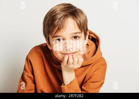 Studio di un bel ragazzo di 10 anni con capelli biondi, con cappuccio marrone, posato su sfondo bianco Foto Stock