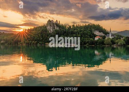 l'effetto sunburst mentre il sole tramonta dietro il castello di bled crea una splendida scena di castello di bled e della chiesa di san martino riflessa nel lago di bled Foto Stock