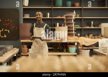 Giovane vasaio africano con una tavoletta sorridente in un grande laboratorio di ceramica Foto Stock