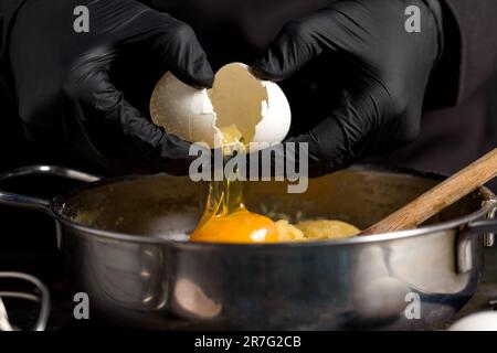 Uno chef professionista in una giacca nera rompe l'uovo in un recipiente di metallo per mescolare l'impasto. Il concetto del processo di cottura Foto Stock