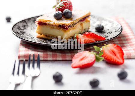 Un pezzo di torta Napoleone con semi di papavero e ciliegie, decorato con fragole, mirtilli su sfondo chiaro. Primo piano Foto Stock