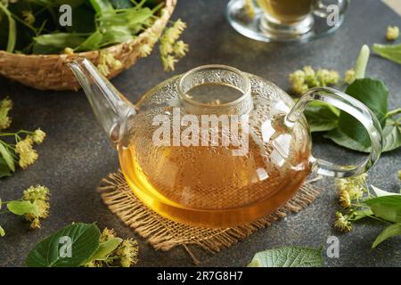 Una teiera di vetro di tisana con fiori di tiglio freschi e foglie su un tavolo Foto Stock
