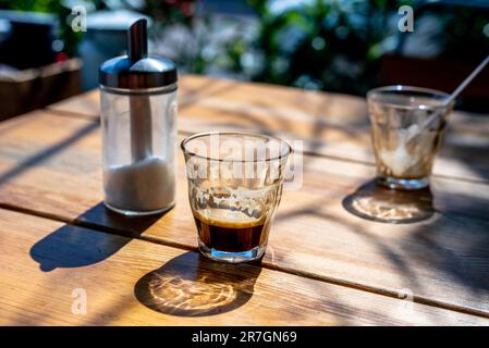 Tazza di caffè espresso nero caldo, un bicchiere vuoto e zucchero sul tavolo nel bar di strada nelle ore del tramonto. Foto Stock
