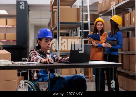 Dipendente asiatico del magazzino in sedia a rotelle che esegue l'ottimizzazione dell'inventario delle merci sul laptop. Operatore di spedizione del servizio postale che lavora sul computer alla scrivania in un magazzino accessibile ai disabili Foto Stock