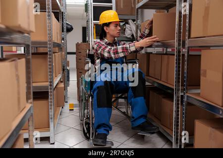 Giovane operaio asiatico in carrozzina che prende il pacco dal ripiano. Commissionatore di magazzino con disabilità fisica cercando scatola di cartone durante il lavoro in magazzino Foto Stock