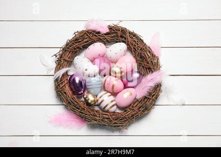 Uova di Pasqua decorate festosamente, corona di viti e piume su un tavolo di legno bianco, vista dall'alto Foto Stock