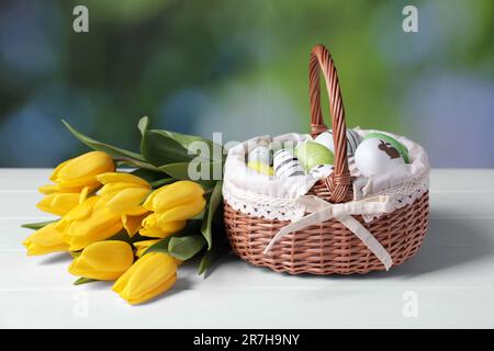 Cestino di vimini con uova di Pasqua decorate in modo festivo e bellissimi tulipani su un tavolo di legno bianco Foto Stock