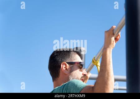 uomo bearded con gli occhiali da sole visto da dietro allenare la schiena facendo pull-up su un barbell con una fascia elastica fitness in una palestra all'aperto. Foto Stock