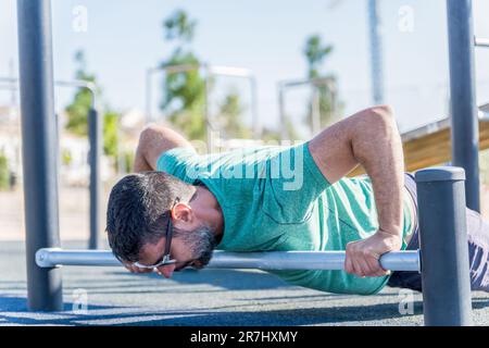 uomo visto in profilo con barba e occhiali da sole che fanno push-up su un bar sul pavimento Foto Stock