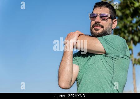 vista frontale di un bel bearded uomo con occhiali da sole che allunga i muscoli in un campo con un cielo blu sullo sfondo. Foto Stock