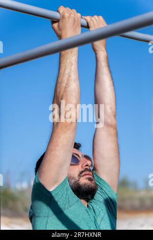 uomo bearded con gli occhiali da sole visto in profilo allenando la schiena facendo pull-up su un barbell in una palestra all'aperto. Foto Stock