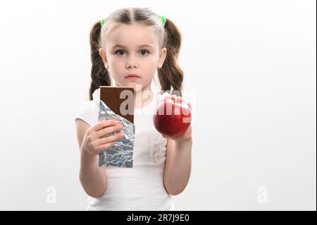 Ritratto di una bambina su sfondo bianco, sceglie il cioccolato su una mela, cibo sano vs cibo spazzatura. Foto Stock