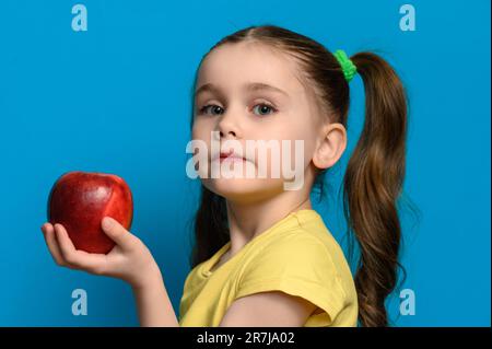 La ragazza si trova in posizione di profilo su uno sfondo blu e tiene una grande mela rossa, spazio copia e pappe per bambini in una mano. Foto Stock