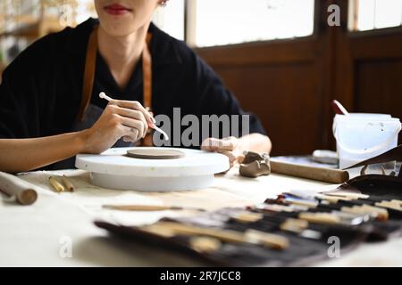 Vista ritagliata del giovane uomo che decora la ceramica prima di cottura in officina. Arte, attività, artigianato e concetto di hobby Foto Stock