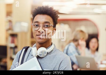 Ritratto di sorridente giovane afro-americano ragazzo studente con capelli ricci indossando occhiali e cuffie intorno al collo in piedi in biblioteca campus Foto Stock