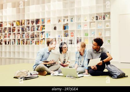 Gruppo di giovani interrazziali e penosi studenti seduti tra documenti e dispositivi sul piano della biblioteca e discutendo idee per un progetto comune Foto Stock