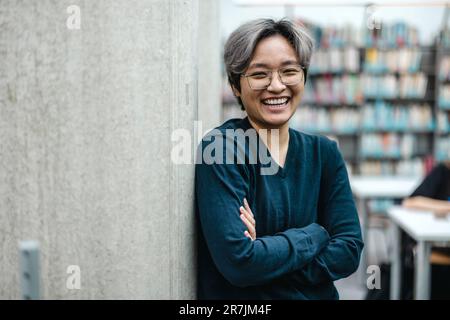 Ritratto di sorridente studente asiatico in piedi con le braccia incrociate in biblioteca del college Foto Stock