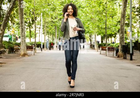 Donna sorridente che cammina in città usando il telefono e prendendo un caffè. Foto Stock