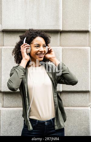 Giovane donna sorridente mentre usa le cuffie per strada. Foto Stock